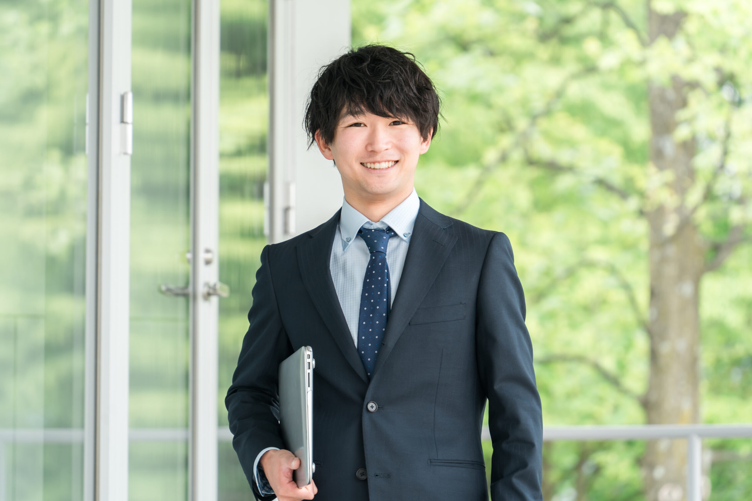 写真：爽やかなグリーンをバックに微笑む若いスーツ姿の男性